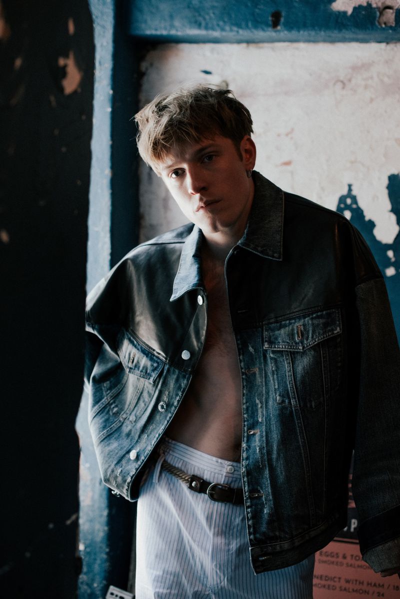 Young adult poses in a denim jacket with blue and white striped shorts against a textured wall indoors, medium shot, eye-level.