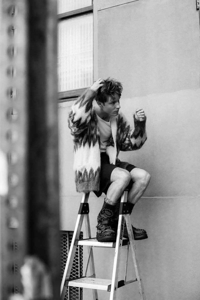 Young man poses on a ladder in an urban setting wearing a patterned sweater, shorts, and boots in this black and white lifestyle portrait.