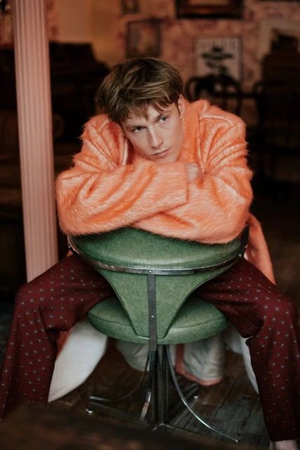 A young man with brown hair sits on a green chair in an indoor setting, wearing a fuzzy orange robe and patterned burgundy pants.