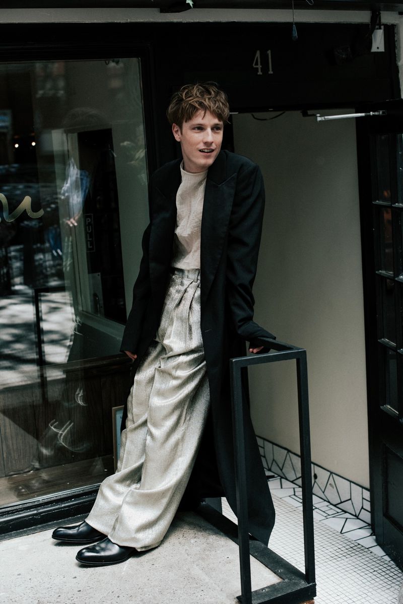 Young adult male fashion model poses outside a building. He wears wide-legged silver pants, a cream top, and a long black jacket.