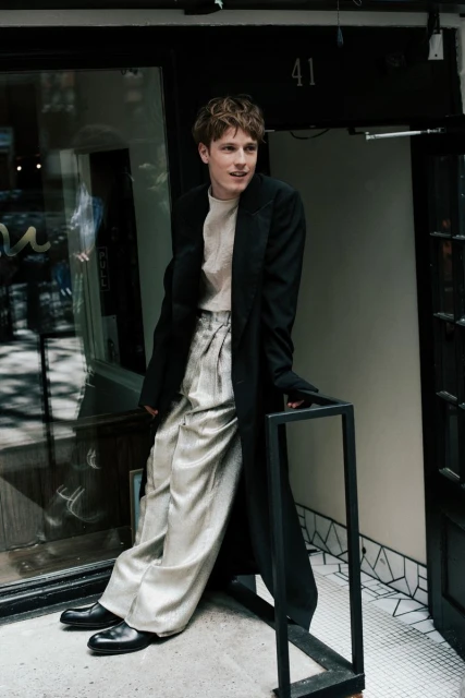 Young adult male fashion model poses outside a building. He wears wide-legged silver pants, a cream top, and a long black jacket.