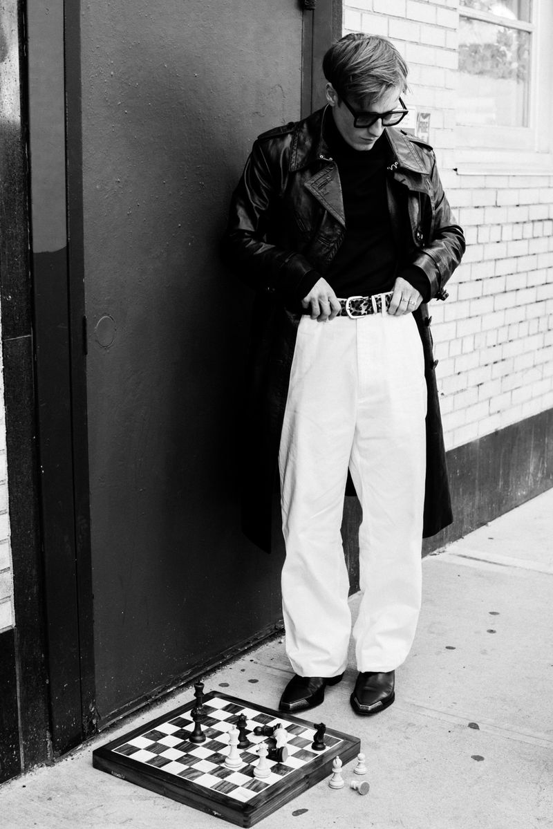 A full shot captures a man in a leather trench coat standing by a door with a chessboard on the ground in black and white.