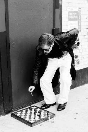 Black and white photo of a man playing chess outside on a city street, wearing sunglasses and a leather jacket, placing a chess piece.