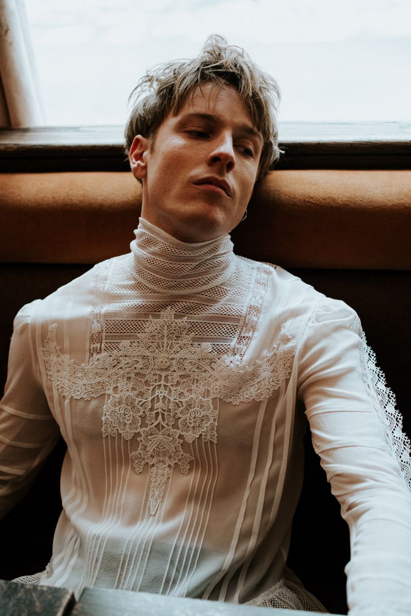 Close-up portrait of a young adult male in a white, lace-trimmed top, posing indoors with a window, capturing a fashion or editorial vibe.