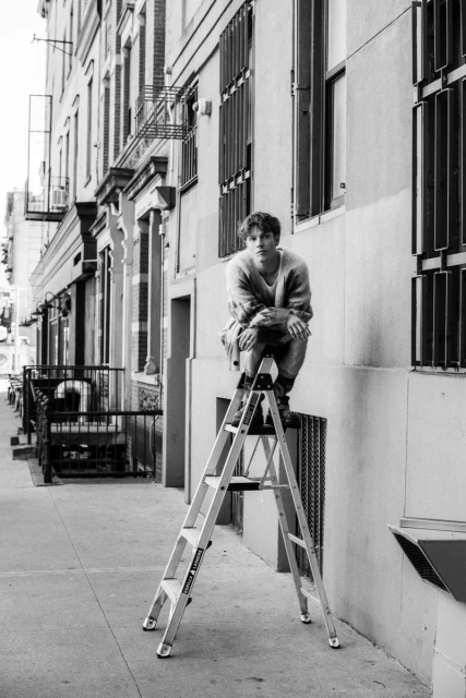 Black and white shot of adult male sitting on top of aluminum ladder on sidewalk in urban environment with building facade in the background.