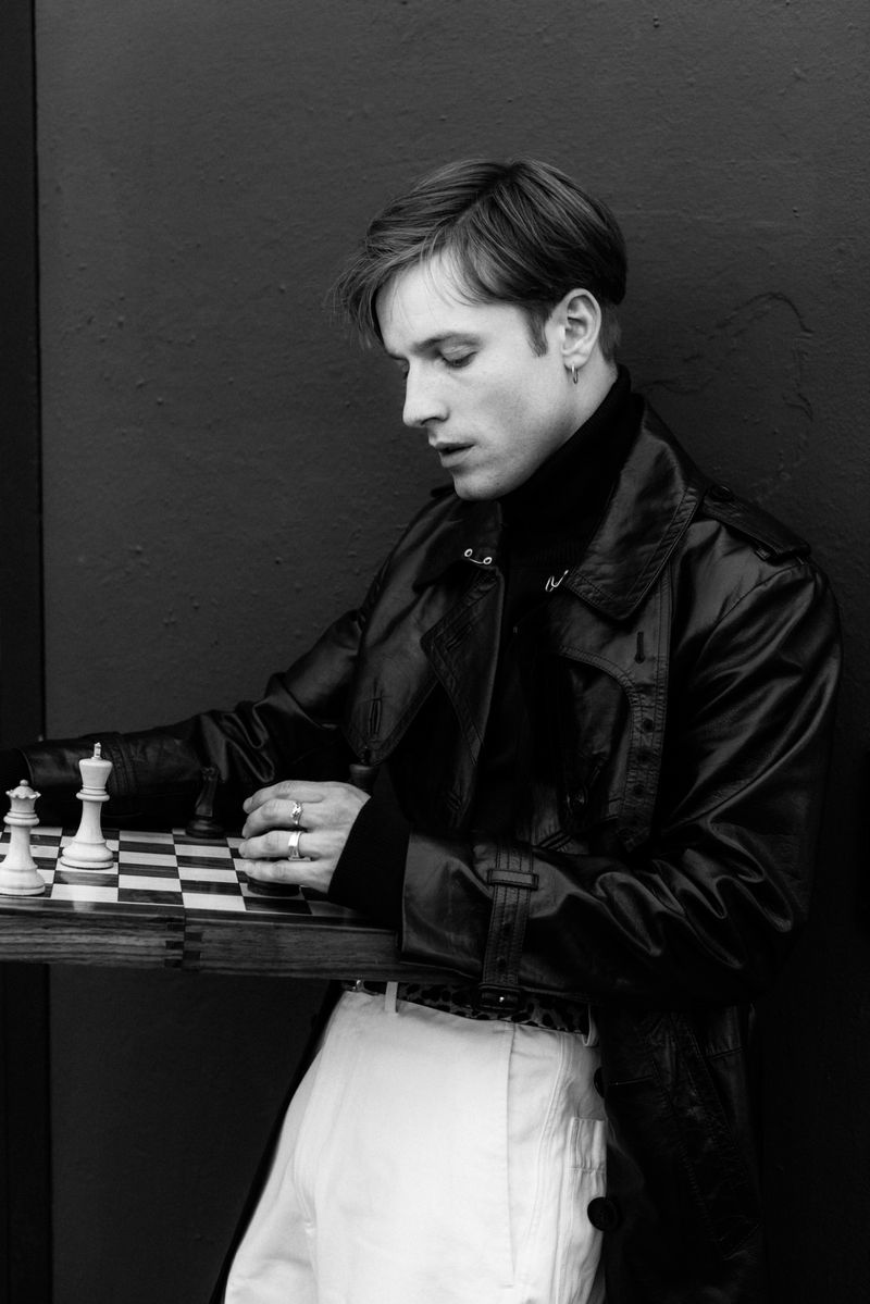 Black and white portrait of a young man in a leather coat playing chess indoors, showcasing fashion and interior elements.