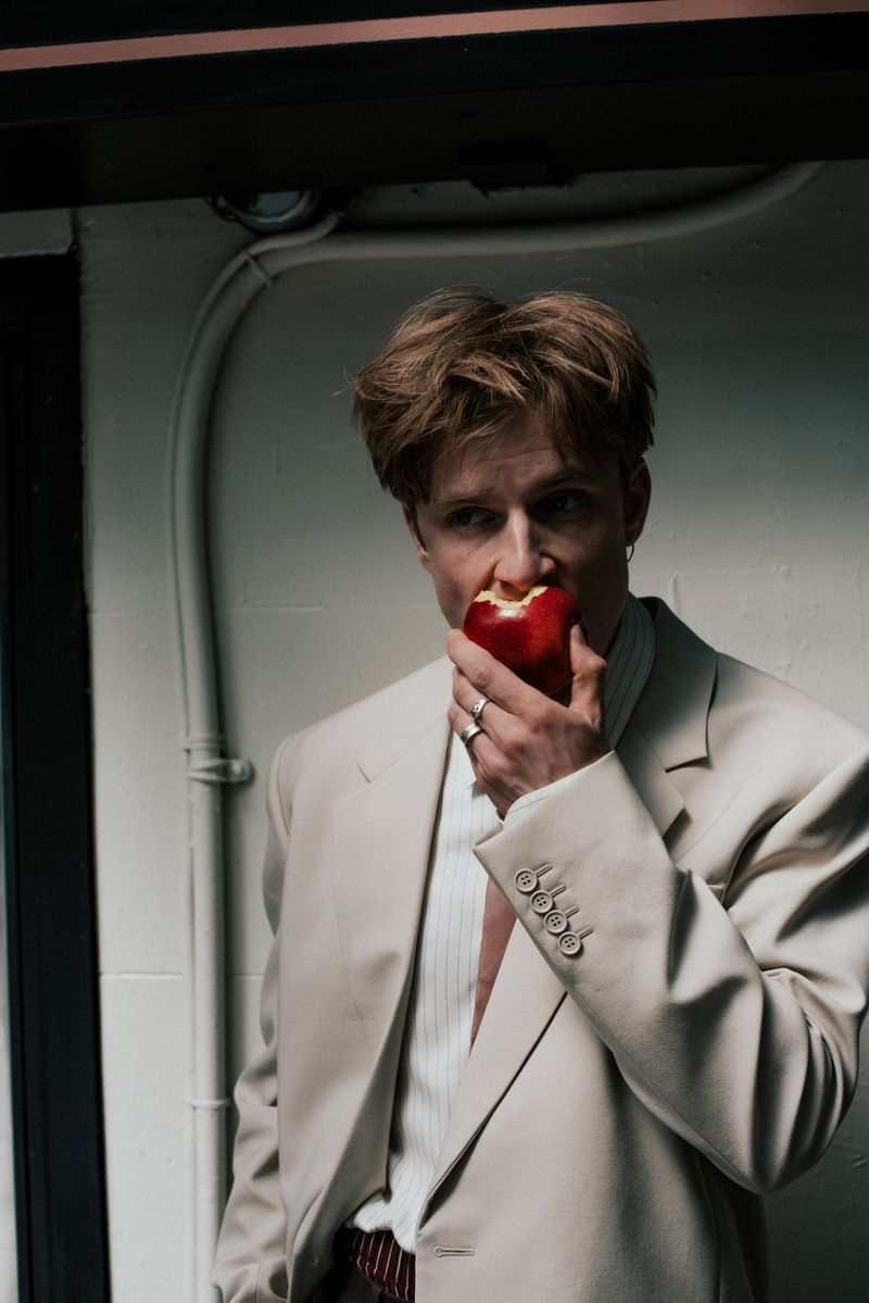 Louis Hofmann wearing an oversized beige tailored suit blazer while biting into a red apple against a white wall.