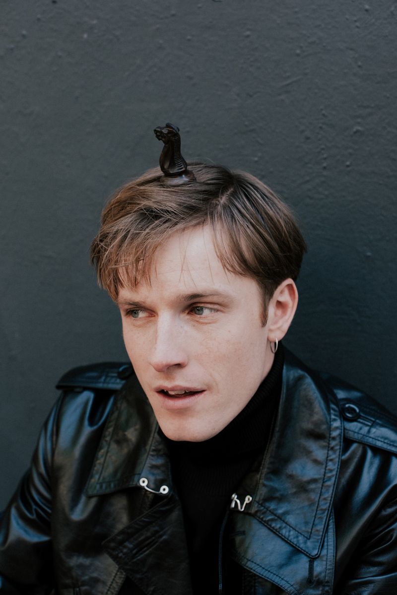 Portrait of a fair-skinned adult male wearing a black leather jacket and black turtleneck with a black chess piece on his head.
