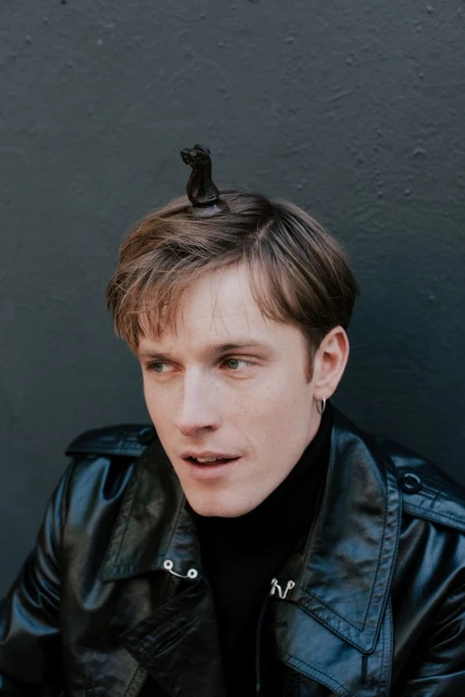 Portrait of a fair-skinned adult male wearing a black leather jacket and black turtleneck with a black chess piece on his head.