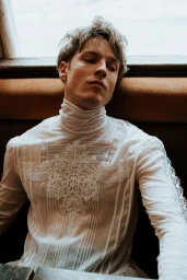 Actor Louis Hofmann wearing a sheer high-neck white lace blouse while leaning back against a brown booth seat.