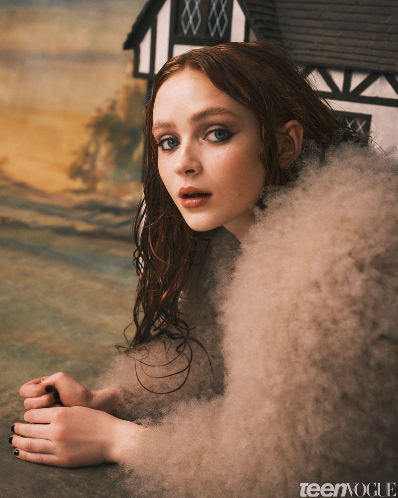 Close-up portrait of a young woman with auburn hair, wearing a cream-colored, fluffy jacket, posing in front of a rustic, illustrated backdrop.