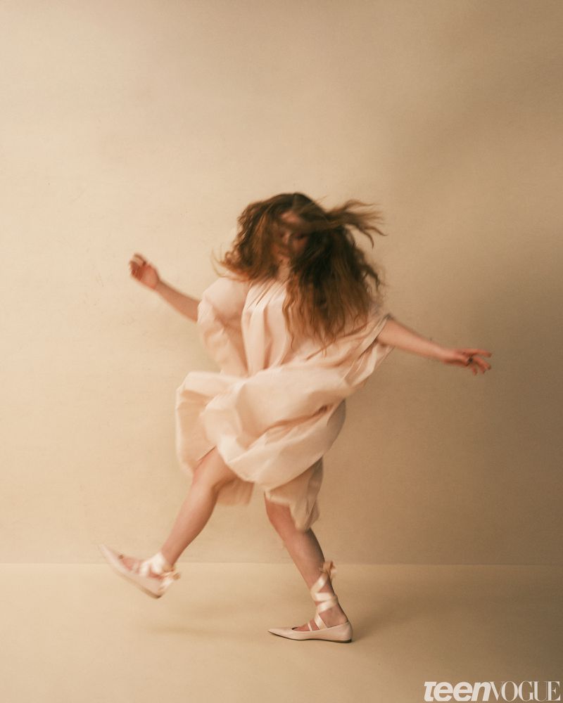 A woman in a light pink dress and ballet shoes leaps against a pale beige background in a fashion-editorial shot with soft studio lighting.