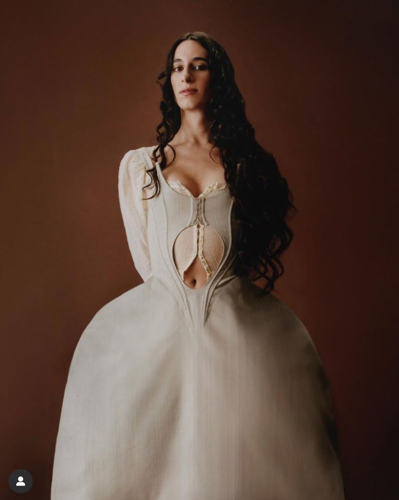Woman posing in studio in a dress with a unique bodice cutout and a wide skirt. Long dark hair and a brown background.