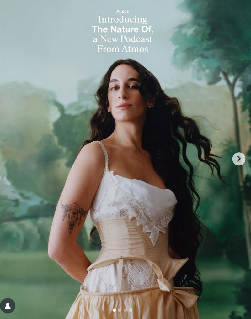 Portrait of woman with long dark hair wearing a corset in front of an outdoor backdrop with trees and painted clouds.