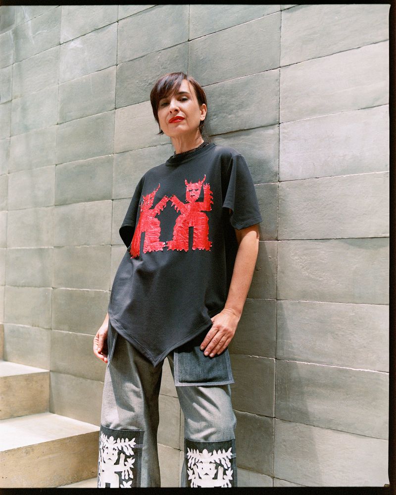 A woman with short dark hair stands near a textured wall, wearing a black t-shirt with red designs and patterned grey and white denim pants.