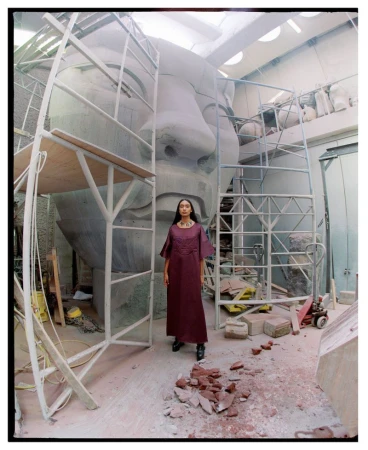 A woman in a maroon dress stands in an industrial space next to a partially constructed, oversized face sculpture under scaffolding.