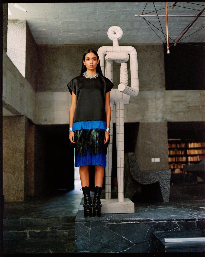 Woman in modern fashion poses in a brutalist interior beside a modern block sculpture. The space features geometric patterns and textures.