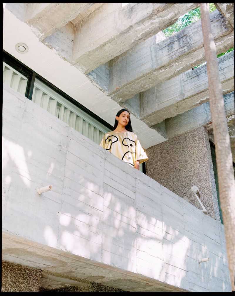 A young adult female poses on a concrete balcony with a building of concrete beams and a tree in the background, outdoors.