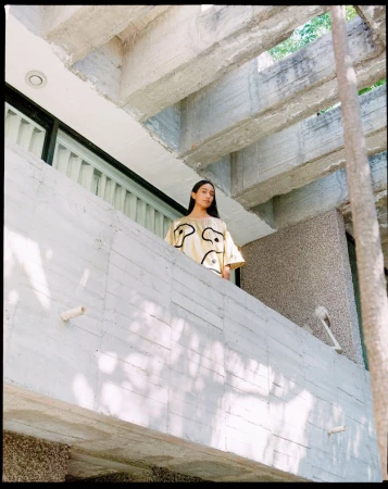 A young adult female poses on a concrete balcony with a building of concrete beams and a tree in the background, outdoors.