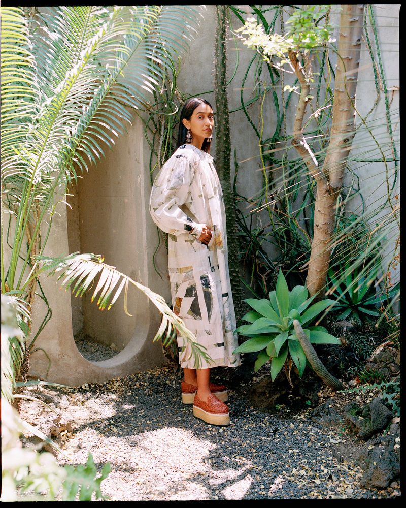 Woman poses in a patterned dress and platform shoes in a courtyard with tropical plants and a stone wall, fashion editorial style.