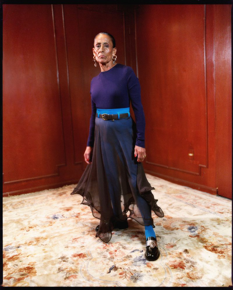 Mature woman in a blue ribbed knit top and layered sheer skirt standing on an ornate floral rug against wood-paneled walls.