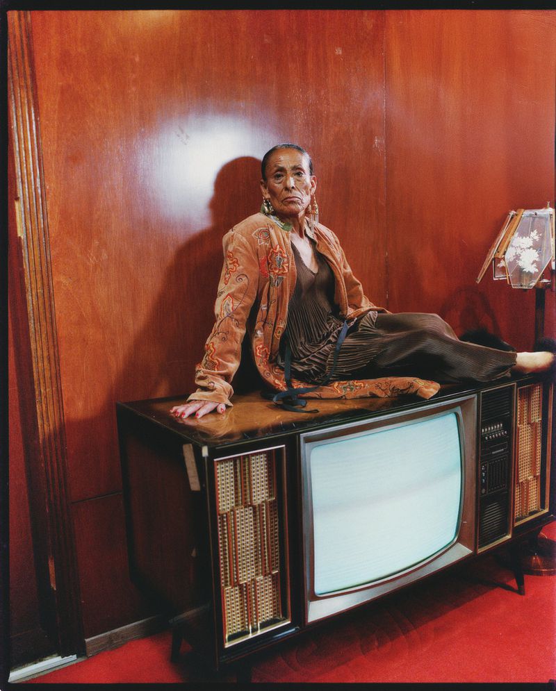 Older Black woman in patterned jacket poses on vintage television in a room with wood paneling and red carpet, creating a retro aesthetic.