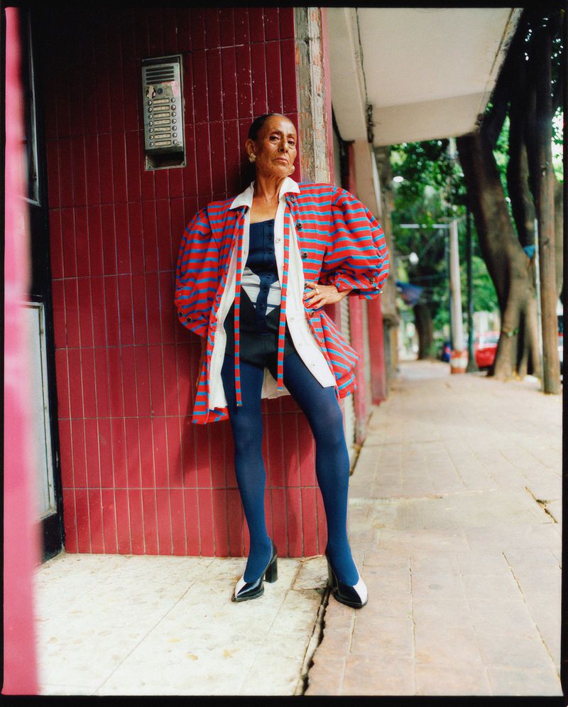 Mature adult female in a striped jacket and blue tights poses outdoors against a red brick wall on a city sidewalk next to green trees.