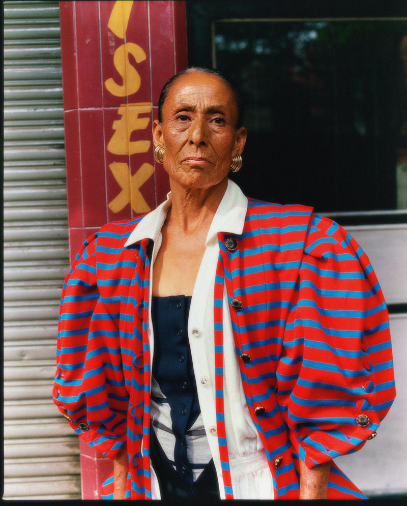 Portrait of a woman with gold hoop earrings wearing a red and blue striped jacket, posing in front of a red tile wall with yellow lettering.