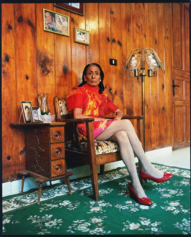 Elderly woman sitting in an armchair, indoors. She wears a red dress and red shoes. A wooden table and lamp are behind her.