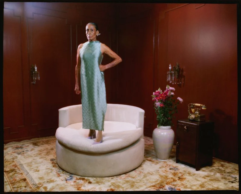 Elegant older woman stands on a cream-colored round chair in a seafoam-green dress with dark red walls and patterned carpet in the background.