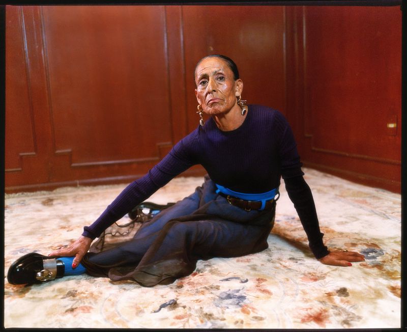Elderly woman sits cross-legged on floral rug in violet top, slate skirt with bright blue accents, statement earrings, and black shoes.