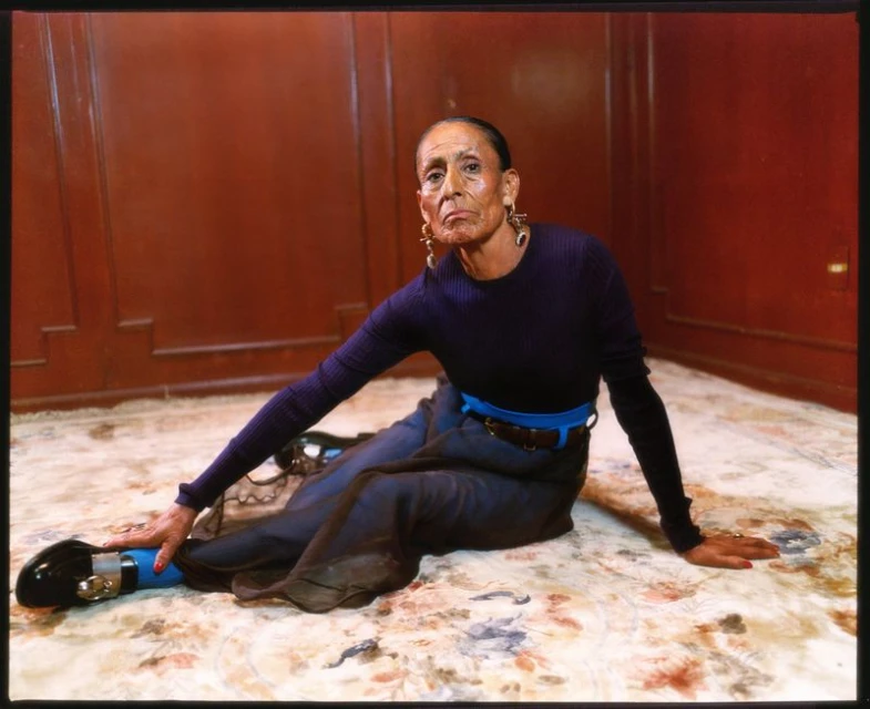 Elderly woman sits cross-legged on floral rug in violet top, slate skirt with bright blue accents, statement earrings, and black shoes.