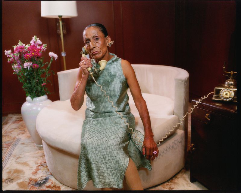 Elegant woman in a dress sits in a chair, holding a vintage telephone receiver, with a vase of pink flowers and furniture nearby.