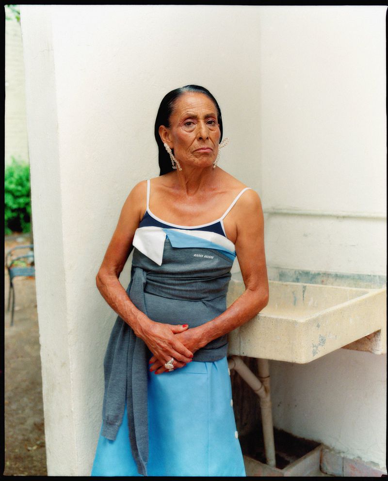 An older woman with dark hair stands indoors wearing a light blue skirt and layered top with large earrings, in a fashion portrait.