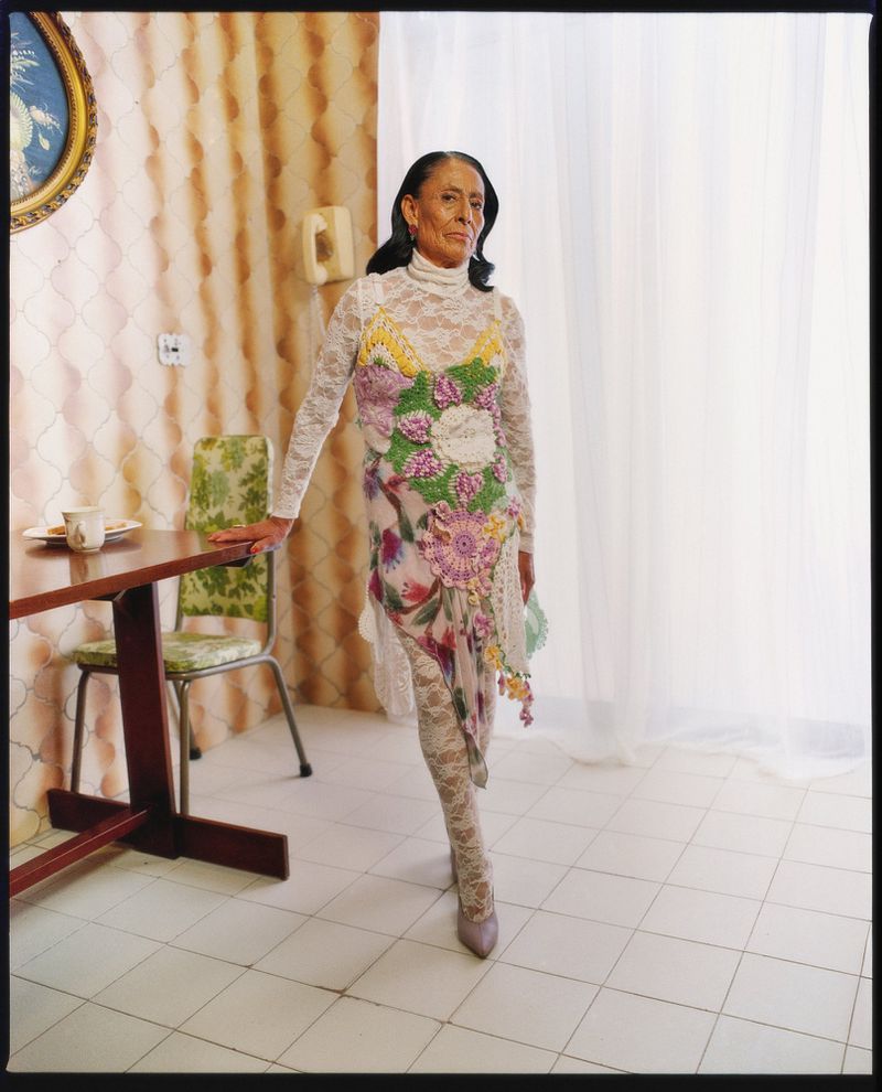Mature woman poses in a floral dress with a lace overlay in an interior with patterned wallpaper, a table, and a white curtain.
