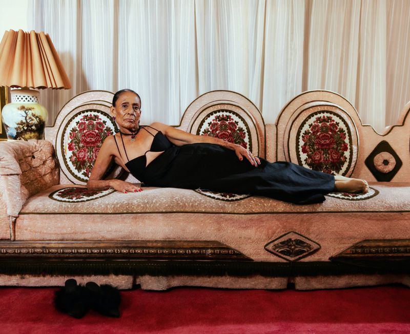 Elegant older woman in a black dress reclines on a pink floral couch in an indoor setting with white curtains and dark carpet.