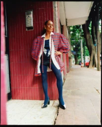 Mature woman in a red and blue striped puff-sleeve coat and blue tights posing against a red tiled wall on a city street.