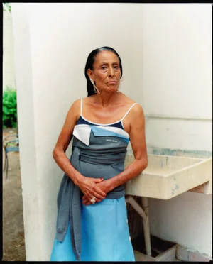 Elderly woman wearing a grey strap top and blue skirt posing outdoors next to a stone sink for Blanc Magazine