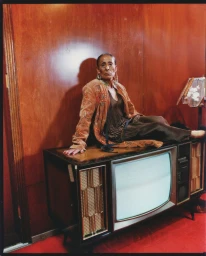 Elderly woman in an embroidered caramel jacket seated on top of a vintage console television against wood-paneled walls.