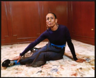 Elderly woman sitting on a floral rug in a dark wood-paneled room wearing a navy sweater, blue belt, and black loafers.
