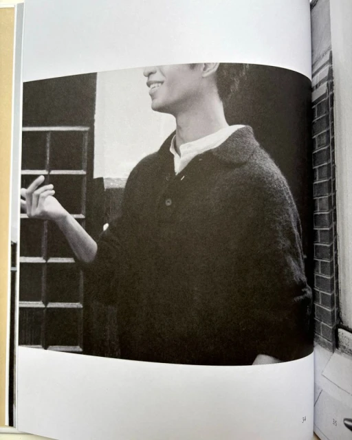 Monochromatic image of an adult male in a buttoned sweater, standing near a window and a brick wall, captured in a medium shot.
