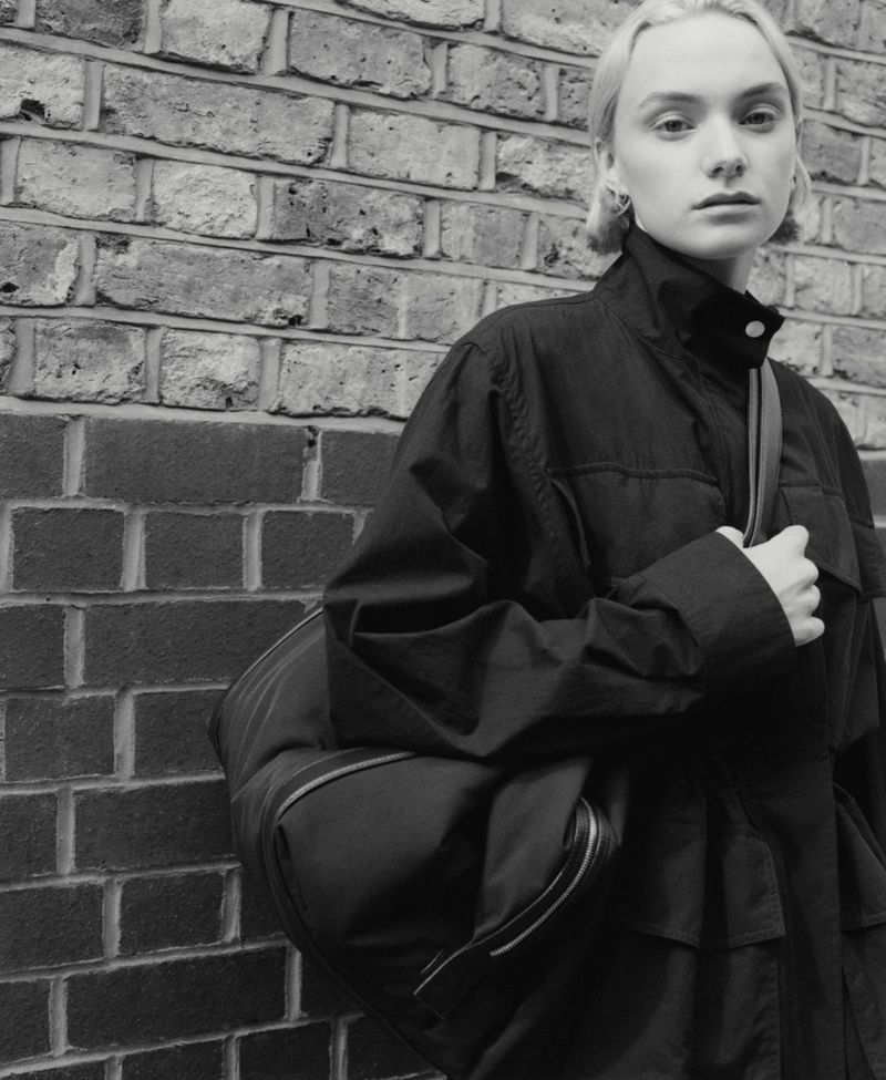 Monochromatic portrait of a young adult woman with short blonde hair in a dark jacket with a bag slung over her shoulder.