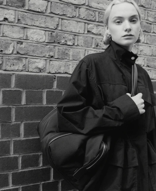 Monochromatic portrait of a young adult woman with short blonde hair in a dark jacket with a bag slung over her shoulder.