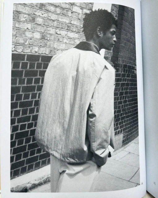 Monochrome image of a man wearing a jacket against a brick wall, creating an urban fashion editorial style photograph.