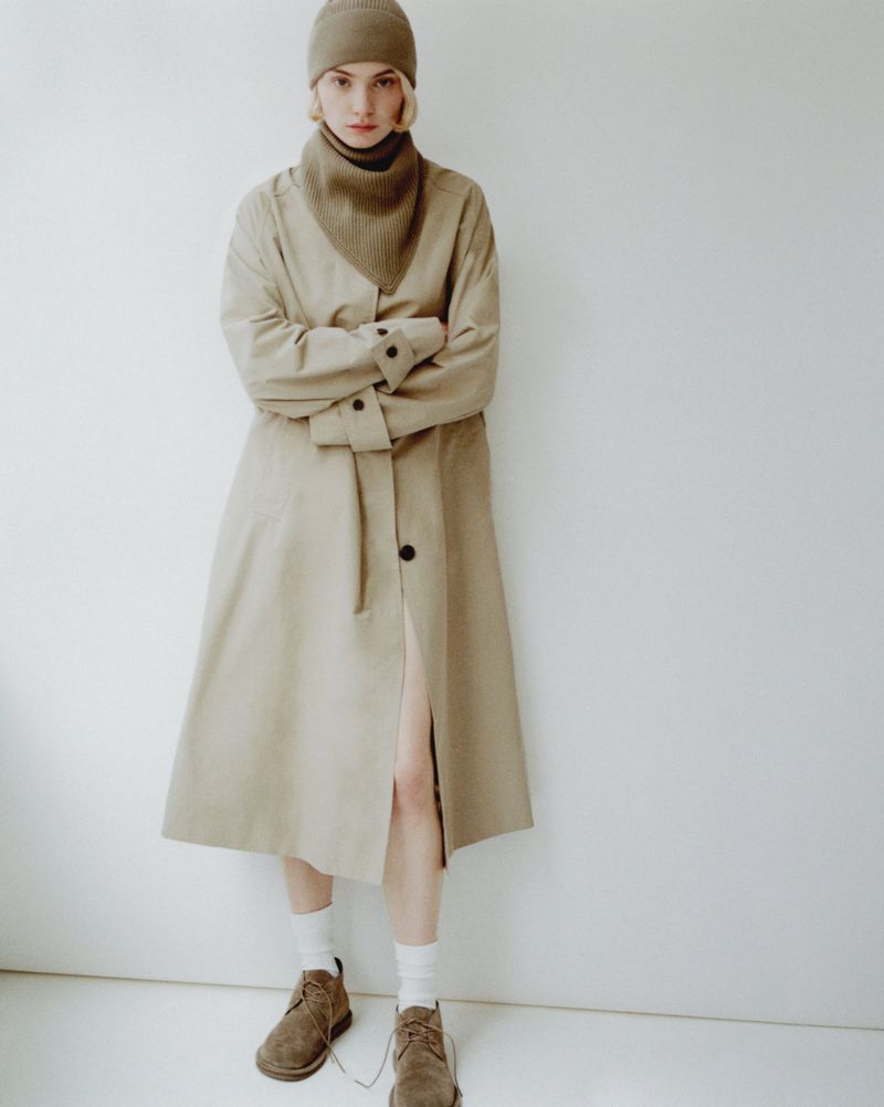 A woman in a beige coat, turtleneck, beanie, and brown shoes stands in front of a white wall in a fashion shot.