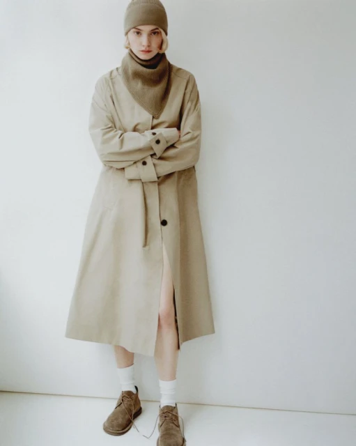 A woman in a beige coat, turtleneck, beanie, and brown shoes stands in front of a white wall in a fashion shot.