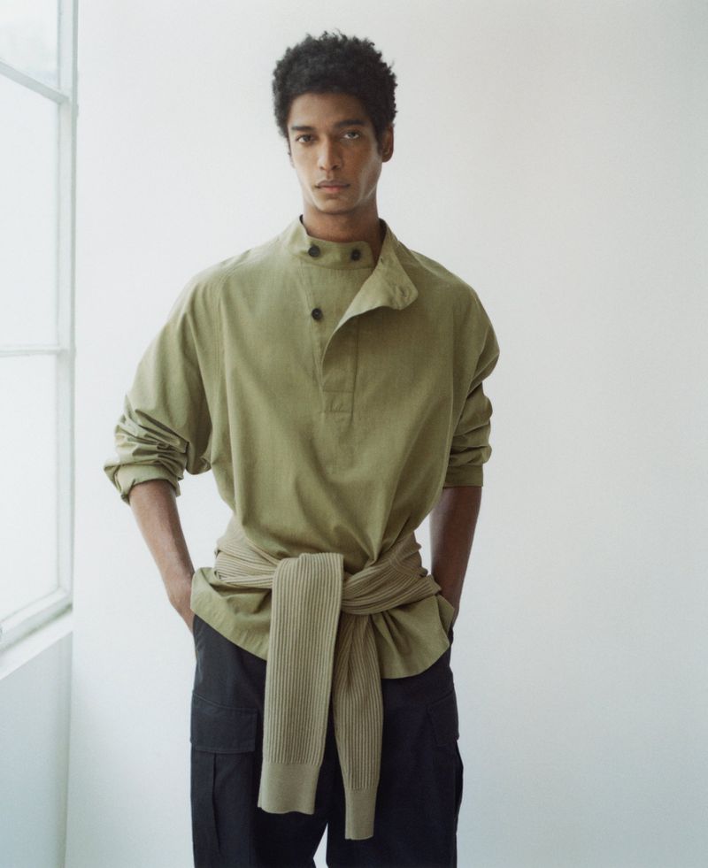 A tan-skinned male fashion model poses in front of a white wall in a green shirt with black cargo pants and a sweater tied at the waist.