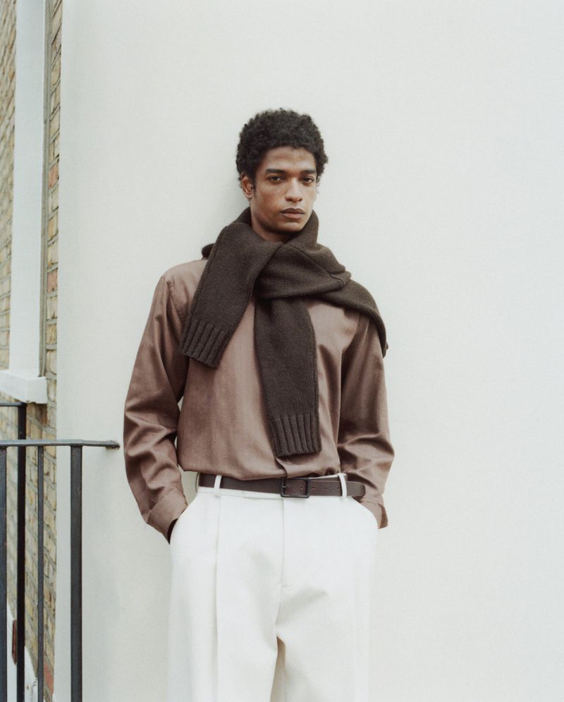 Portrait of a young adult male wearing a brown long-sleeved shirt and scarf with white pants, posing against an off-white wall.