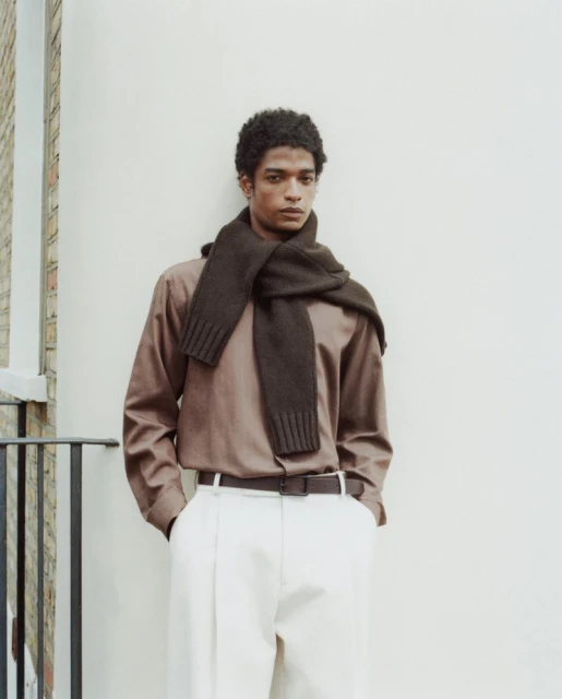 Portrait of a young adult male wearing a brown long-sleeved shirt and scarf with white pants, posing against an off-white wall.