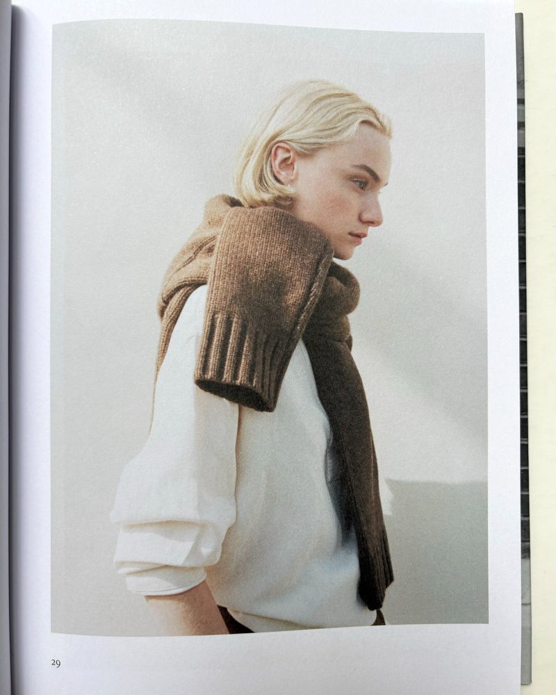 Blonde adult poses in a soft light wearing a white long sleeve shirt and a brown knit scarf draped over the shoulders and chest.
